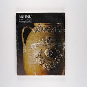 Könnte beinhalten: Ein Buchcover für Brunk Auctions, das eine große, braune, keramische Kanne mit aufwendigen Schnitzereien zeigt. Das Buchcover ist schwarz mit weißem Text, der "BRUNK AUCTIONS" und "November 13-14, 2019" lautet.
