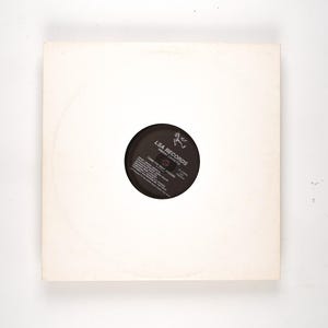 May include: A vinyl record in a white sleeve. The record label is black with white text and the LSA RECORDS logo. The sleeve is square with a circular cutout revealing the record label. The record is centered in the sleeve.
