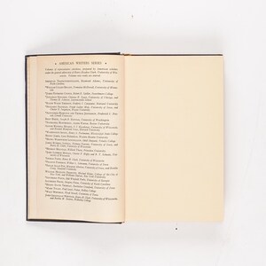 May include: An open book with a dark cover, displaying text on the left page and a blank cream-colored page on the right. The text is a list of names under the title "AMERICAN WRITERS SERIES". The book is set against a white background.