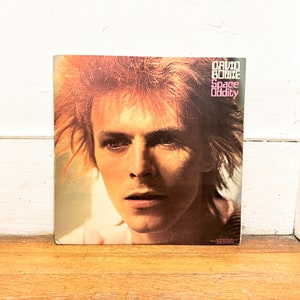 May include: A vintage vinyl record cover featuring a close-up portrait of David Bowie with a red-orange hair style. The album title "Space Oddity" is printed in pink letters on a black background.