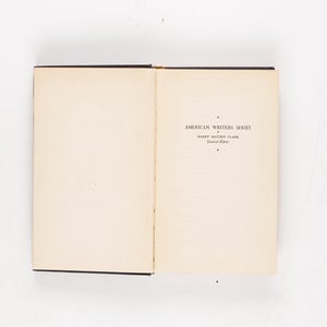 May include: Open book with text reading "AMERICAN WRITERS SERIES" and "HARRY HAYDEN CLARK". The book has a black spine and the pages are off-white. The book is open on a white surface.