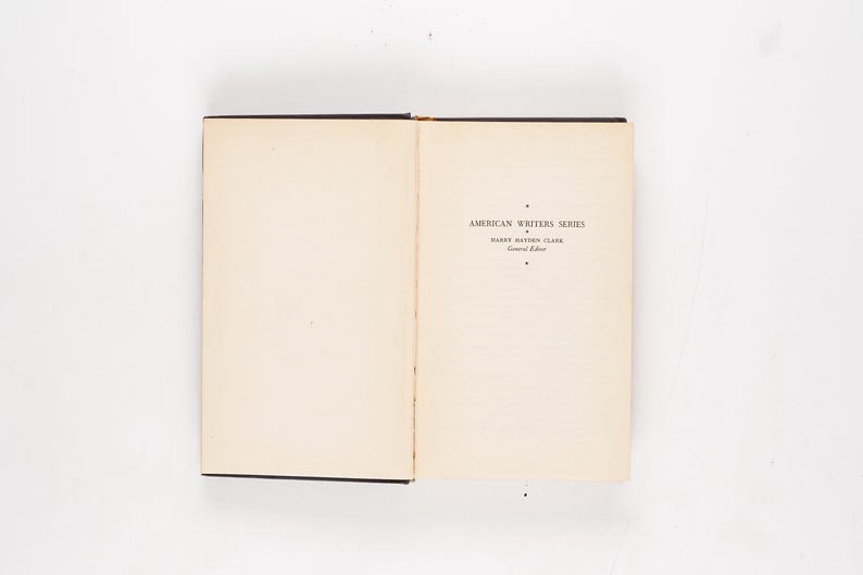 May include: Open book with text reading "AMERICAN WRITERS SERIES" and "HARRY HAYDEN CLARK". The book has a black spine and the pages are off-white. The book is open on a white surface.