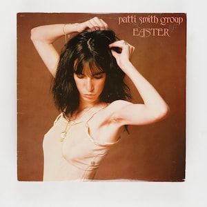 Puede incluir: Portada del álbum "Easter" de Patti Smith Group. La imagen presenta a una mujer con cabello oscuro, vistiendo una blusa de color claro, con los brazos levantados. El título del álbum y el nombre del artista están en una fuente estilizada en la parte superior.