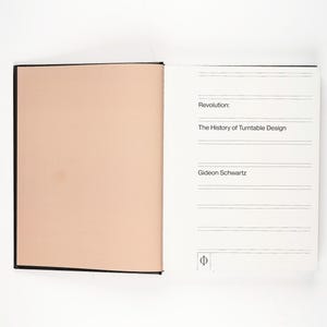May include: An open book with a black spine and cover. The left page is a solid peach color, while the right page has text that reads "Revolution: The History of Turntable Design" and the author's name, Gideon Schwartz.