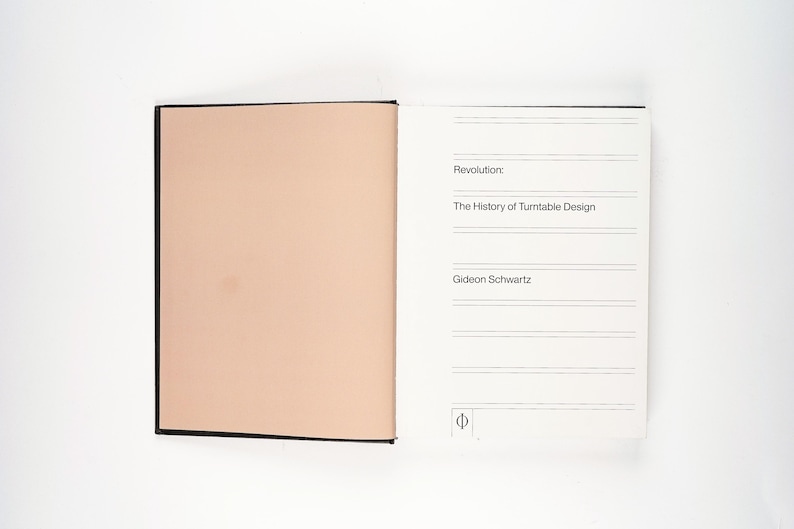 May include: An open book with a black spine and cover. The left page is a solid peach color, while the right page has text that reads "Revolution: The History of Turntable Design" and the author's name, Gideon Schwartz.