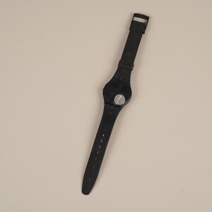May include: Black wristwatch with a round face and a black strap. The watch has a silver-colored dial and a buckle closure. The strap is made of a flexible material and has several holes for adjustment.