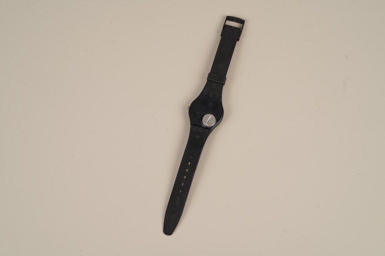 May include: Black wristwatch with a round face and a black strap. The watch has a silver-colored dial and a buckle closure. The strap is made of a flexible material and has several holes for adjustment.
