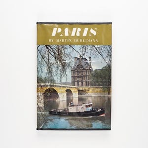 Pode incluir: Uma capa de livro vintage com fundo dourado e o título "PARIS" em texto branco. A capa apresenta uma foto de uma ponte sobre um rio com um barco na água e um edifício no fundo.