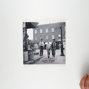 May include: A black and white photo book cover featuring three men walking down a street. The book title is "Cuchi White in Black and White" and the author is "Ezra Stoller, photographer, with an introduction by Lloyd Kramer".