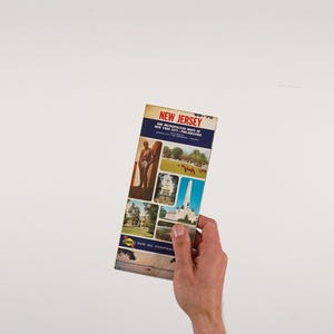 May include: A vintage New Jersey map from 1969-70, held in a hand. The map features a blue and white banner with the state name and a list of cities. Several small images of landmarks and attractions are displayed on the map.