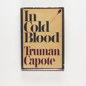 May include: A book cover for "In Cold Blood" by Truman Capote. The title and author's name are in bold black and maroon lettering on a tan background. A red graphic is also visible on the cover.