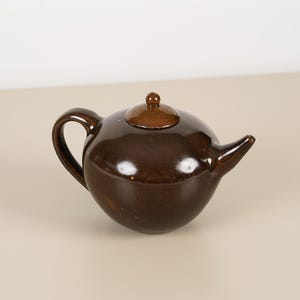 May include: A small, round, brown ceramic teapot with a matching lid and curved handle. The teapot has a glossy finish and a spout for pouring. The design is simple and classic, suitable for serving tea.