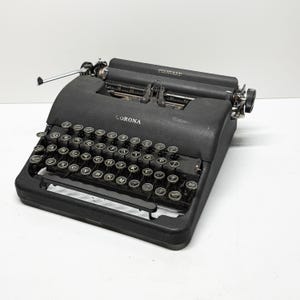以下が含まれることがあります： ヴィンテージの黒いコロナスタンダードタイプライター。タイプライターは、丸いキーとキャリッジリターンレバーを備えたフルキーボードを備えています。「CORONA」の文字が機械の前面に表示されています。用紙ローラーは黒で、機械は白い表面に置かれています。