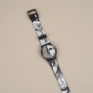 May include: Black and white wristwatch with a patterned band featuring illustrations. The watch face has a black background with a white design. The band is adorned with various black and white images, including clocks and figures.