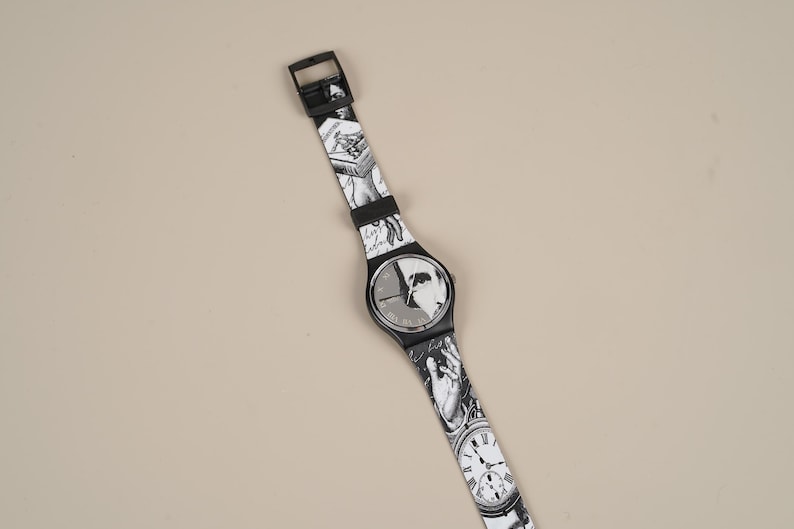 May include: Black and white wristwatch with a patterned band featuring illustrations. The watch face has a black background with a white design. The band is adorned with various black and white images, including clocks and figures.