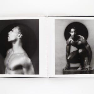 May include: A monochrome photograph displayed across two pages of a book. One image shows a man looking upwards, the other a man crouching. Each image features a dark circular backdrop.