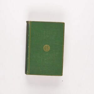 May include: A vintage hardcover book with a green cover and gold accents. The book has a gold emblem in the centre and gold trim around the edges. The spine is black. The book is on a white background.