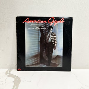 May include: A black vinyl record album cover with the title "American Gigolo" in red and white text. The cover features a man in a striped suit standing in a room with a window. The text "Original Soundtrack Recording" and "Music Composed by Giorgio Moroder" is printed below the title.