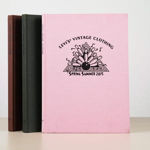 May include: Three hardcover books standing upright. The front book is light pink with black text reading Levi's Vintage Clothing Spring Summer 2015 and a graphic of bowling pins. The middle book is dark green and the back book is dark brown.