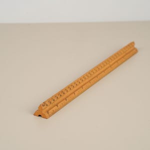May include: A light brown triangular architectural scale ruler made of wood. The ruler features three distinct sides with printed black measurement markings and numerical increments along the edges, designed for drafting and technical drawing tasks.