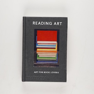 May include: A hardcover book titled "READING ART" with a dark gray cover. The cover features a rectangular window showcasing a stack of colorful books. The text "ART FOR BOOK LOVERS" is below the window. The book is on a white background.