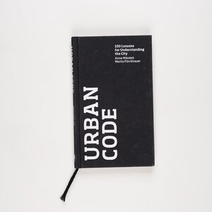 Op de afbeelding: Een zwart boek met de titel "URBAN CODE" in witte hoofdletters. Het boek heeft ook de titel "100 Lessen voor het begrijpen van de stad" en de namen van de auteurs. Een zwart bladwijzer is bevestigd.