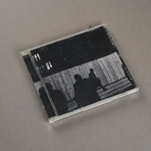 May include: A CD case with a black and white image of silhouetted figures in front of a wooden fence. The image has a grainy texture, suggesting a vintage or artistic aesthetic. The case is clear, and the background is a solid gray.