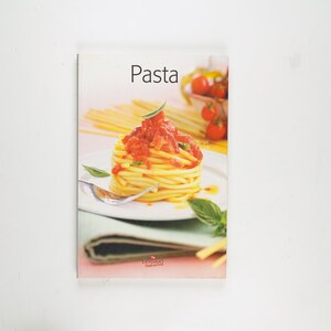 May include: A cookbook titled "Pasta" featuring a vibrant image of spaghetti with tomato sauce, basil, and a fork on a white plate. The cover also shows fresh tomatoes and pasta.