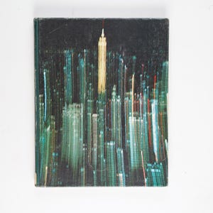Könnte beinhalten: Ein Vintage-Buchcover mit einer nächtlichen Stadtlandschaft mit hohen Gebäuden und einem hellen Licht auf dem höchsten Gebäude.