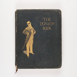 May include: A black book with gold lettering that reads "The Dunlop Book". A gold illustration of a man in a top hat and suit is on the front cover.