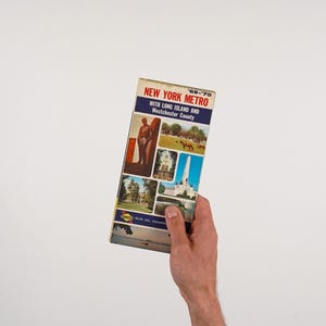 May include: A vintage '69-'70 New York Metro map with Long Island and Westchester County, held by a person. The map features a red, white, and blue color scheme with various images. The Sun Oil Company logo is visible.