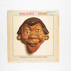 May include: A square vinyl record cover for Pink Floyd Relics featuring a brown, four-eyed face mask with a protruding tongue. The border is cream with a fine dot pattern. Text reads Pink Floyd Relics and A Bizarre Collection of Antiques & Curios.