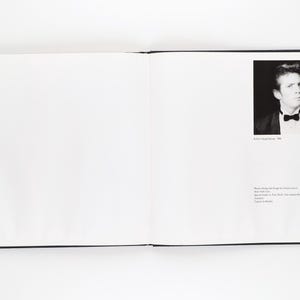 May include: An open book with a black cover. On the right page is a black and white portrait of a man in a tuxedo with a bow tie. Text below the portrait reads "Robert Mapplethorpe, 1986". Further text is present.