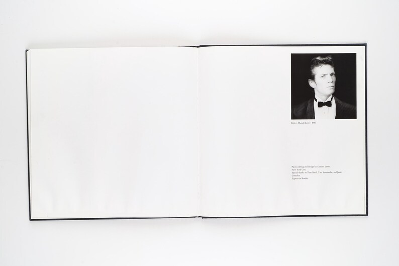 May include: An open book with a black cover. On the right page is a black and white portrait of a man in a tuxedo with a bow tie. Text below the portrait reads "Robert Mapplethorpe, 1986". Further text is present.