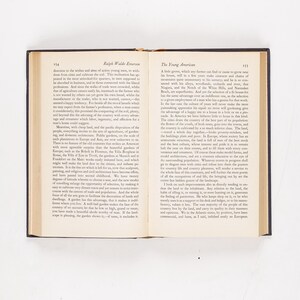 May include: Open book with black cover, displaying text on cream-colored pages. The text includes the title "Ralph Waldo Emerson" and "The Young American." The book is open on a white surface.