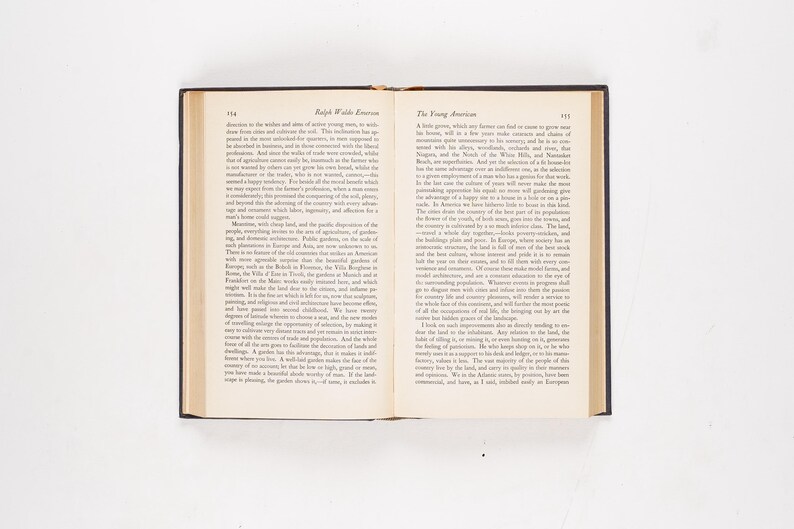 May include: Open book with black cover, displaying text on cream-colored pages. The text includes the title "Ralph Waldo Emerson" and "The Young American." The book is open on a white surface.