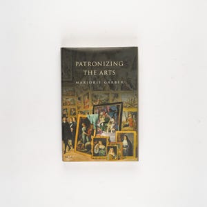 Puede incluir: Un libro titulado "Patronizing the Arts" de Marjorie Garber. La portada presenta un fondo oscuro con una galería de pinturas enmarcadas. El libro está de pie sobre una superficie blanca.