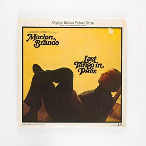 May include: Vintage vinyl record album cover for "Last Tango in Paris" featuring Marlon Brando. The cover has a yellow background with a silhouette of a person reclining. The album title and artist's name are in a stylized font.