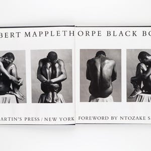 May include: An open book titled "ROBERT MAPPLETHORPE BLACK BOOK" with black and white photographs of a person in various poses. The book is open, displaying four images. The text "ST. MARTIN'S PRESS / NEW YORK FOREWORD BY NTOZAKE SHANGE" is also visible.