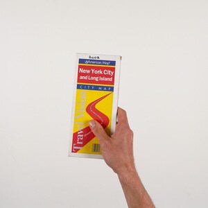 May include: A 2002 New York City and Long Island city map held against a white background. The map has a red and yellow design with the words "Travel Vision" and "City Map" printed on it.