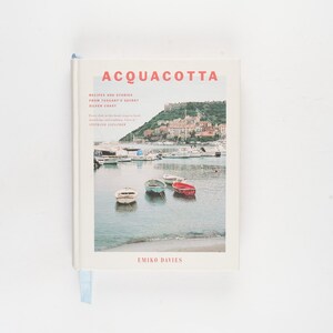 Könnte beinhalten: Ein Hardcover-Buch mit dem Titel "ACQUACOTTA" mit einer Küstenlandschaft auf dem Cover. Das Cover zeigt ein Foto von Booten im Wasser mit einer Stadt im Hintergrund. Das Buch hat ein hellblaues Lesezeichen.