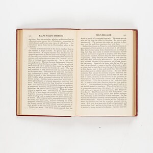 May include: An open book with a red cover and aged pages. The left page displays "Ralph Waldo Emerson" at the top, while the right page reads "Self-Reliance." The book is centered on a white surface.
