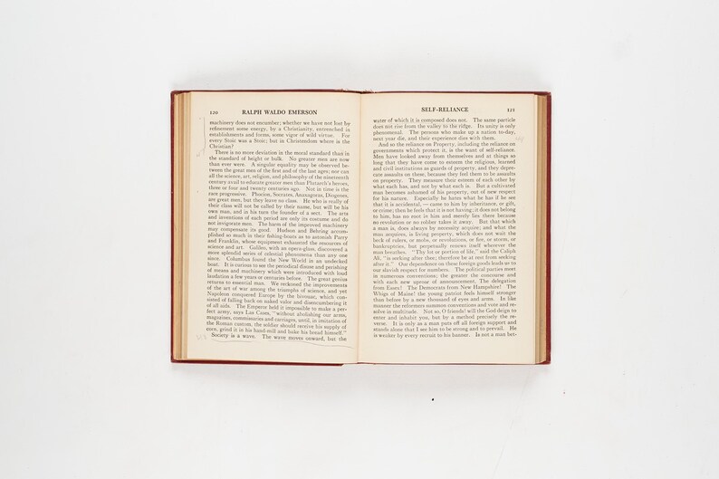 May include: An open book with a red cover and aged pages. The left page displays "Ralph Waldo Emerson" at the top, while the right page reads "Self-Reliance." The book is centered on a white surface.