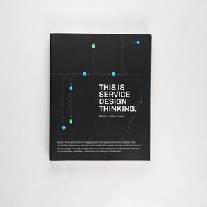 Può includere: Un libro nero intitolato "THIS IS SERVICE DESIGN THINKING". La copertina presenta un diagramma di cerchi blu e rettangoli verdi collegati da linee. Il titolo è bianco, con testo aggiuntivo sotto.