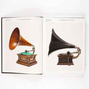 May include: Two antique gramophones are displayed in an open book. One features a wooden horn and a wooden cabinet with a green turntable. The other has a black horn and a wooden cabinet.