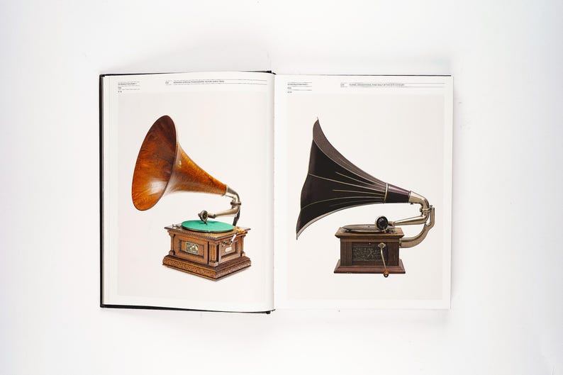 May include: Two antique gramophones are displayed in an open book. One features a wooden horn and a wooden cabinet with a green turntable. The other has a black horn and a wooden cabinet.