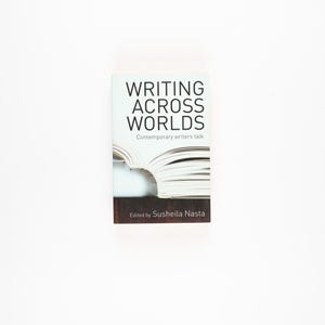 Op de afbeelding: Een boek getiteld "WRITING ACROSS WORLDS" met de ondertitel "Contemporary writers talk" wordt getoond. De omslag toont een gedeeltelijke afbeelding van een open boek met witte pagina's en een donkerbruine basis. De naam van de auteur is ook zichtbaar.
