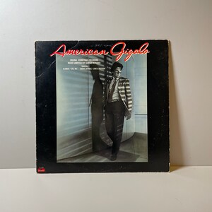 Puede incluir: Una portada de disco de vinilo negro con el título "American Gigolo" en texto rojo y blanco. La portada presenta a un hombre con traje de pie en una habitación con una gran ventana. El texto "Original Soundtrack Recording" está impreso debajo del título.