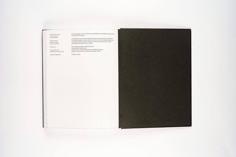 May include: An open book with a black cover and white pages. The left page has text, while the right page is solid black. The book is on a white surface.
