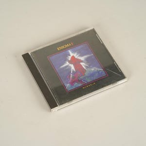 May include: A CD case featuring the album "Enigma I" with a dark cover. The artwork depicts a red figure against a bright, star-like background with the text "ENIGMA I" at the top. The CD case is clear plastic.
